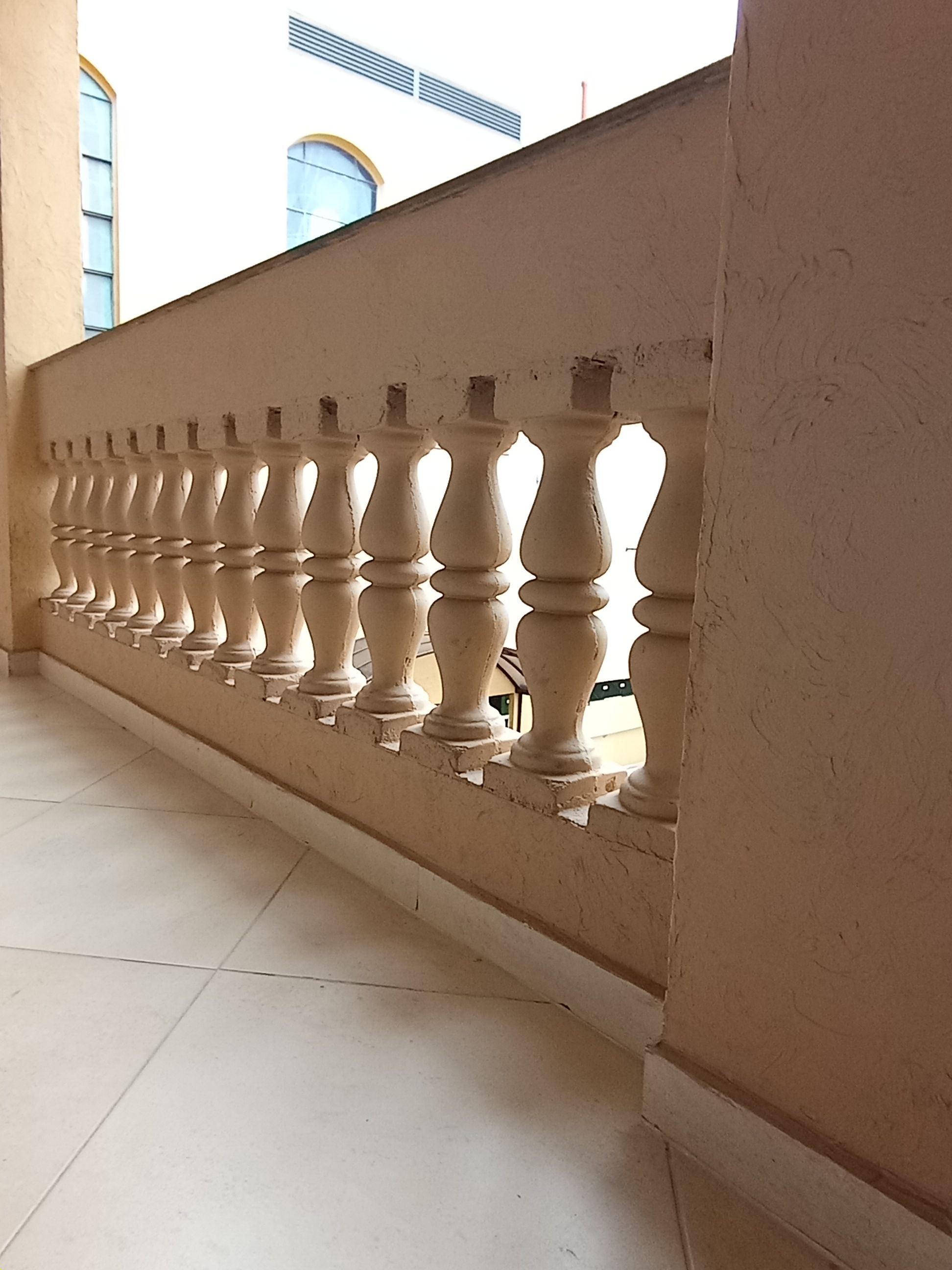 Uma foto em ângulo diagonal e contra-plongé mostra um guarda-corpo clássico com balaústres de concreto bege em uma sacada ou corredor externo. O chão é revestido com pisos cerâmicos claros em formato diagonal, e uma parede de textura grossa na mesma tonalidade bege surge em primeiro plano à direita. Ao fundo, por entre e acima da estrutura, é possível notar partes de um edifício sob a luz do dia.